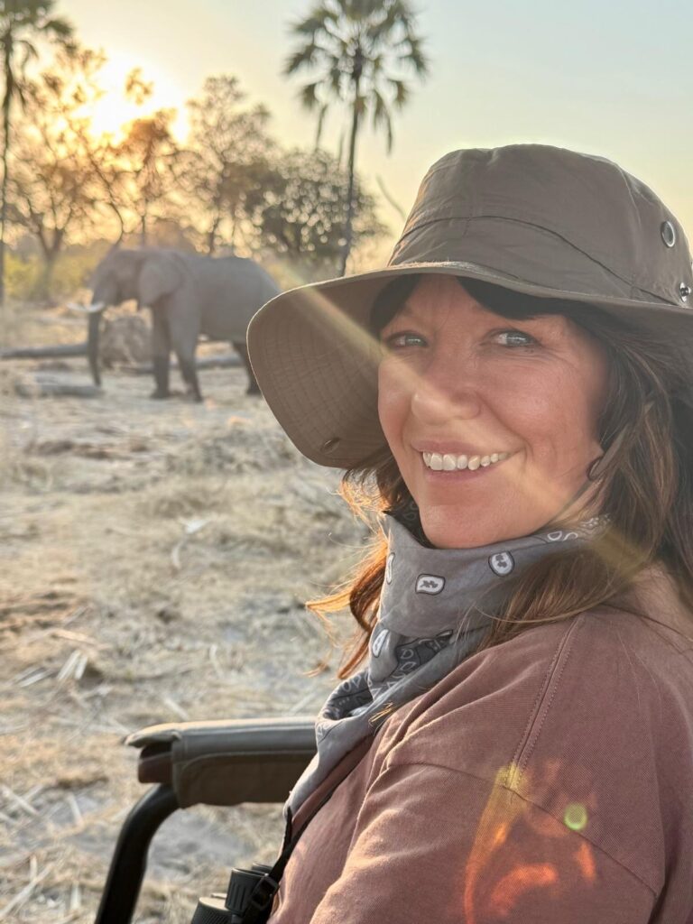 Allison is smiling at the camera, wearing a safari hat with an elephant in the background.