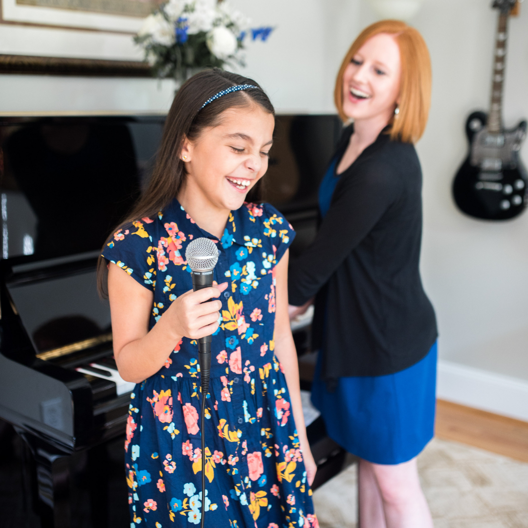 voice student holds a micropone and laughs with her voice teacher as they stand in front of a piano