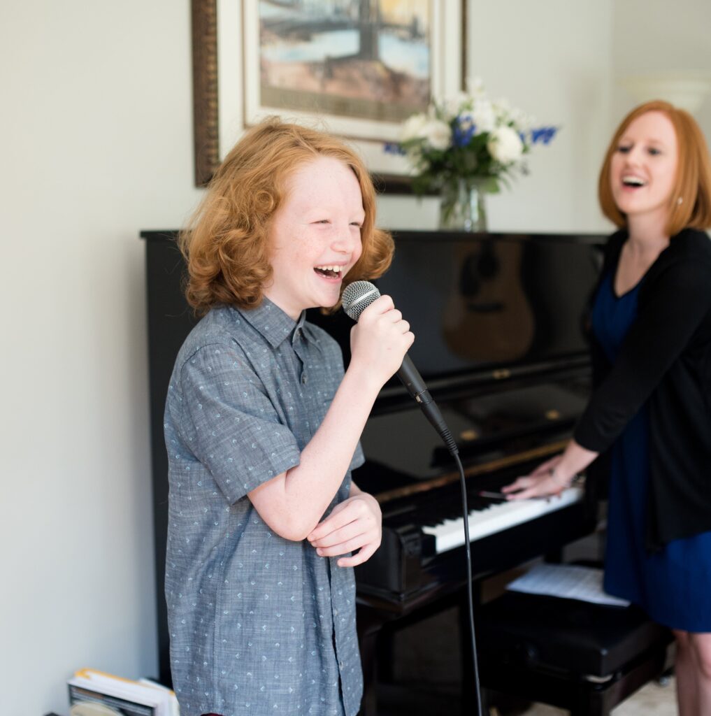 red-headed child sings into a microphone while standing next to a woman with red hair in front of a piano