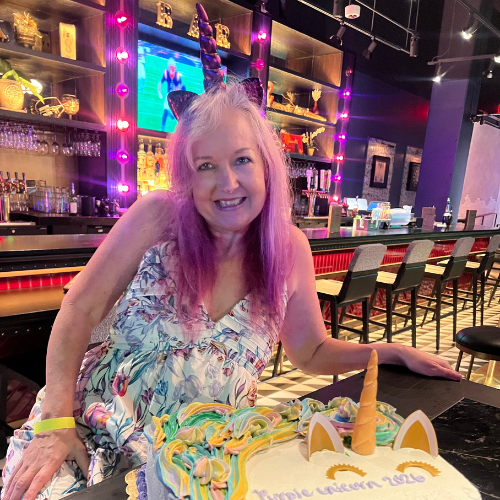 MMM staff member Nita stands in front of a Purple Unicorn cake, while sporting her own purple unicorn horn